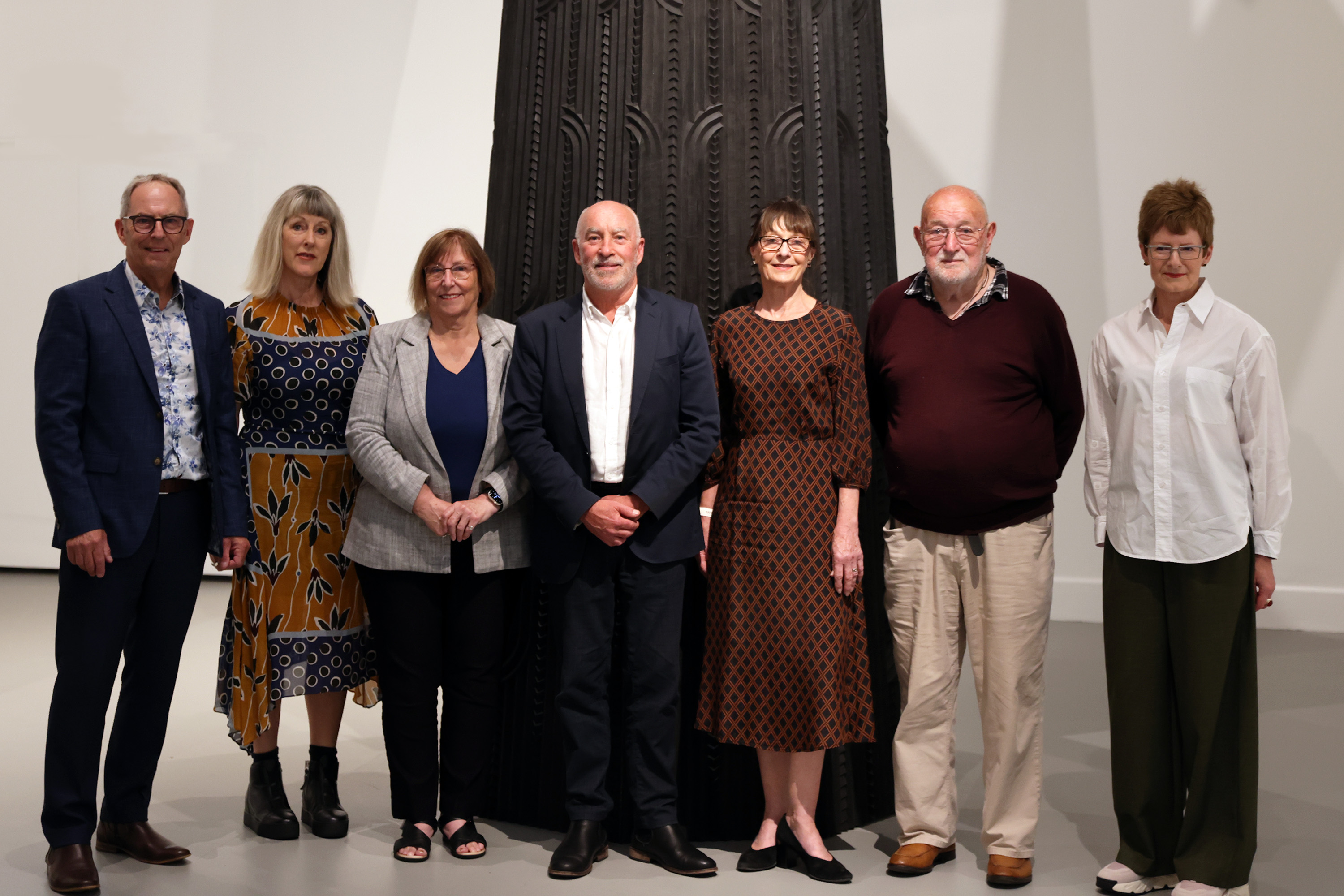 A group of people standing in front of the base of a large black carved sculpture, in a white art gallery space. The sculpture is adorned with fine Māori carvings. 