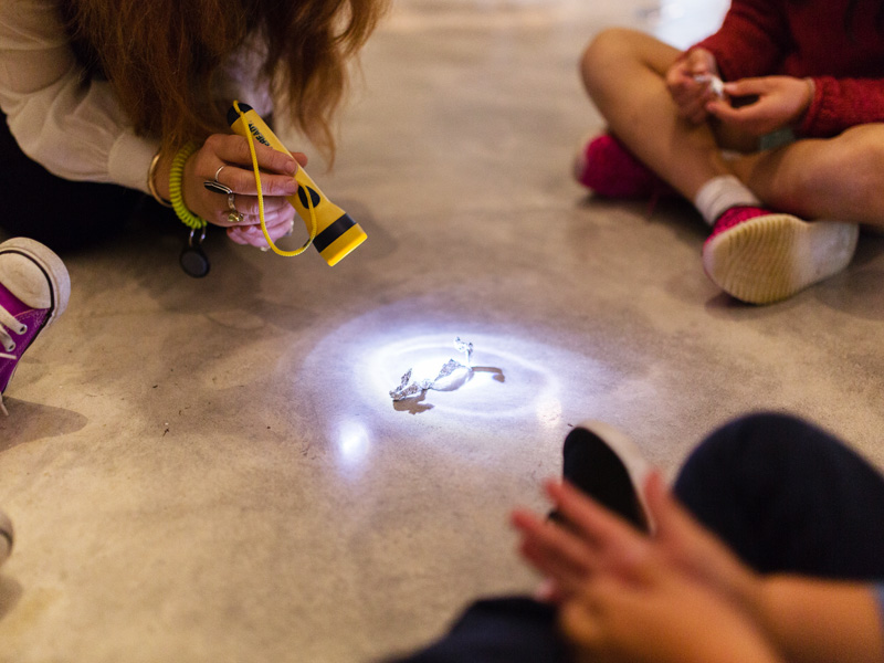 Children sitting on the ground, participating in a science-based art activity involving a torch and shadows