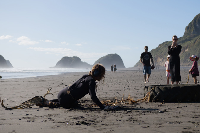 A person, wrapped in woven flax, is sitting on the sand next to a large tractor tyre. They are connected to the tyre by a hand-made flax rope.  